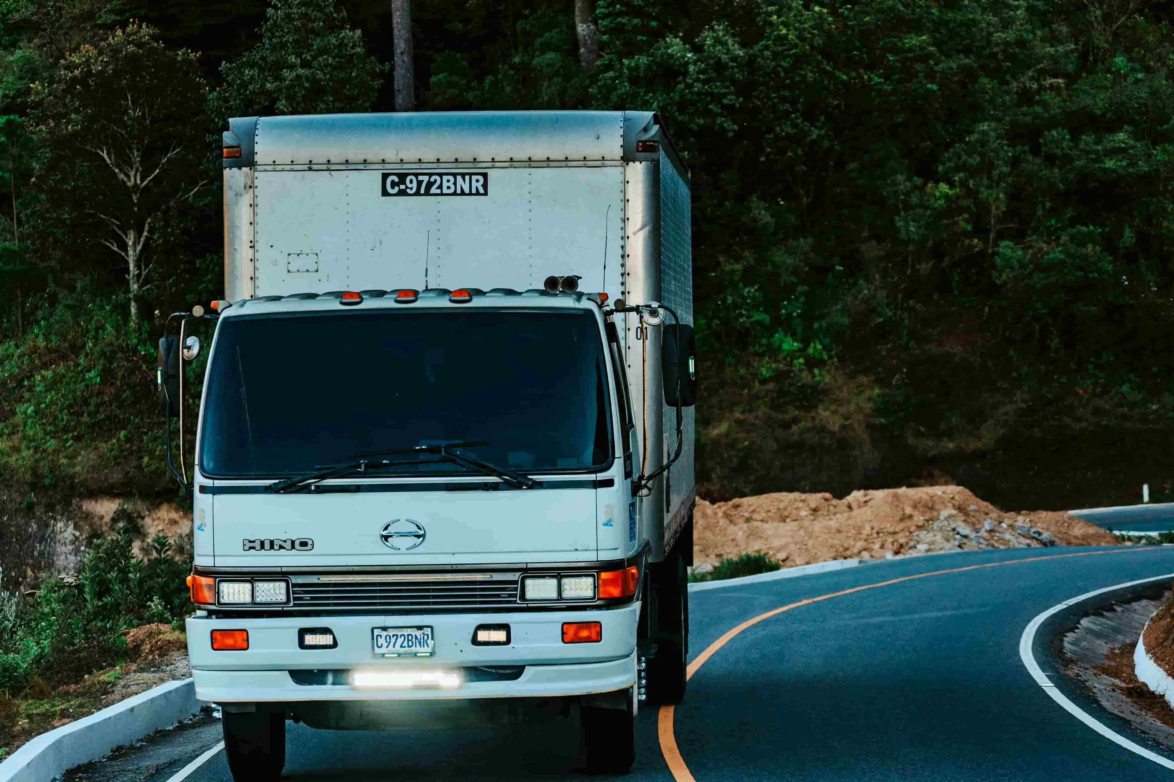 White cab-over straight truck driving a curved two-lane road past piles of excavated dirt