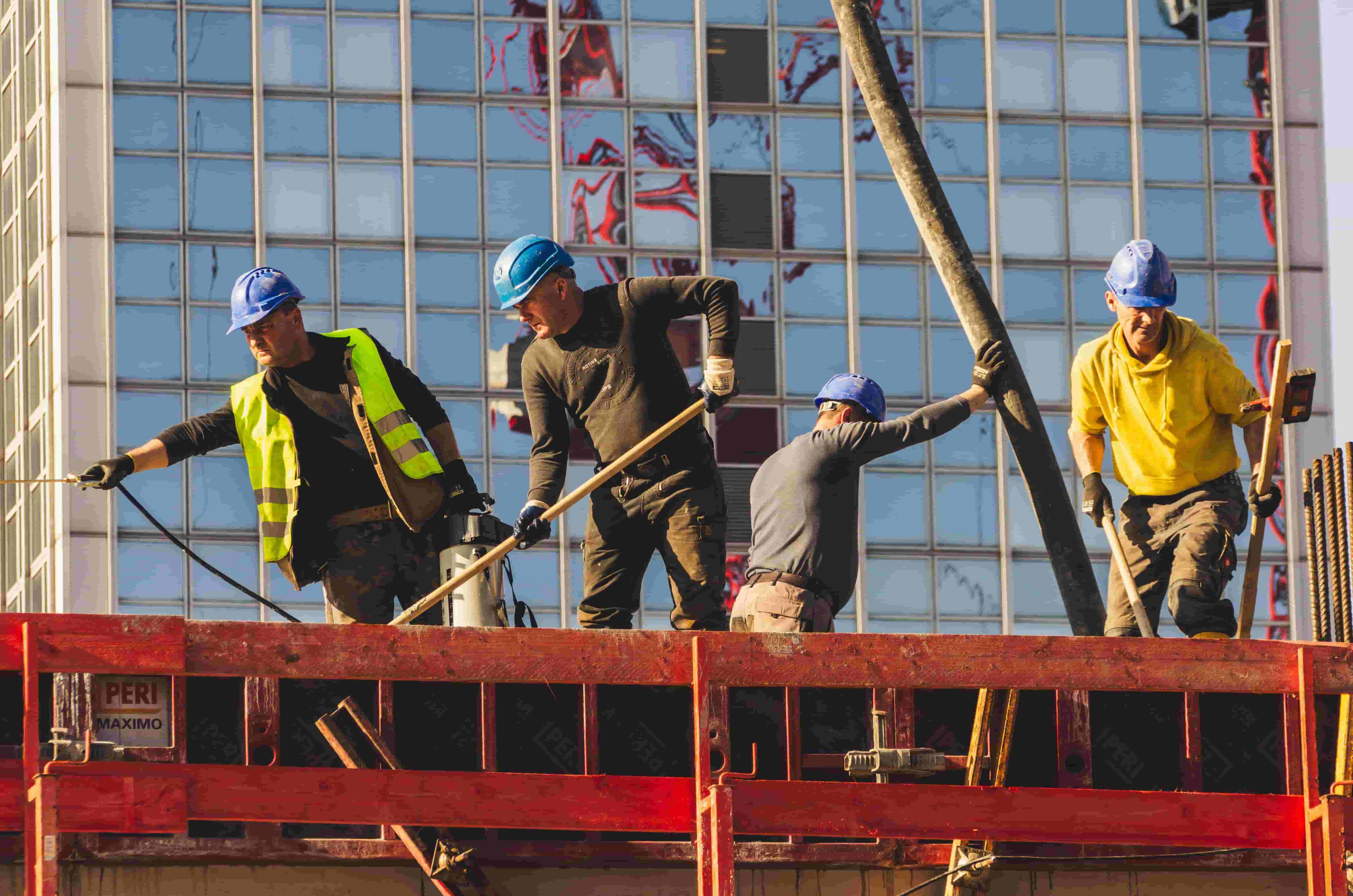 Construction crew in hard hats guiding a concrete pump hose on a building site in front of a glass office tower