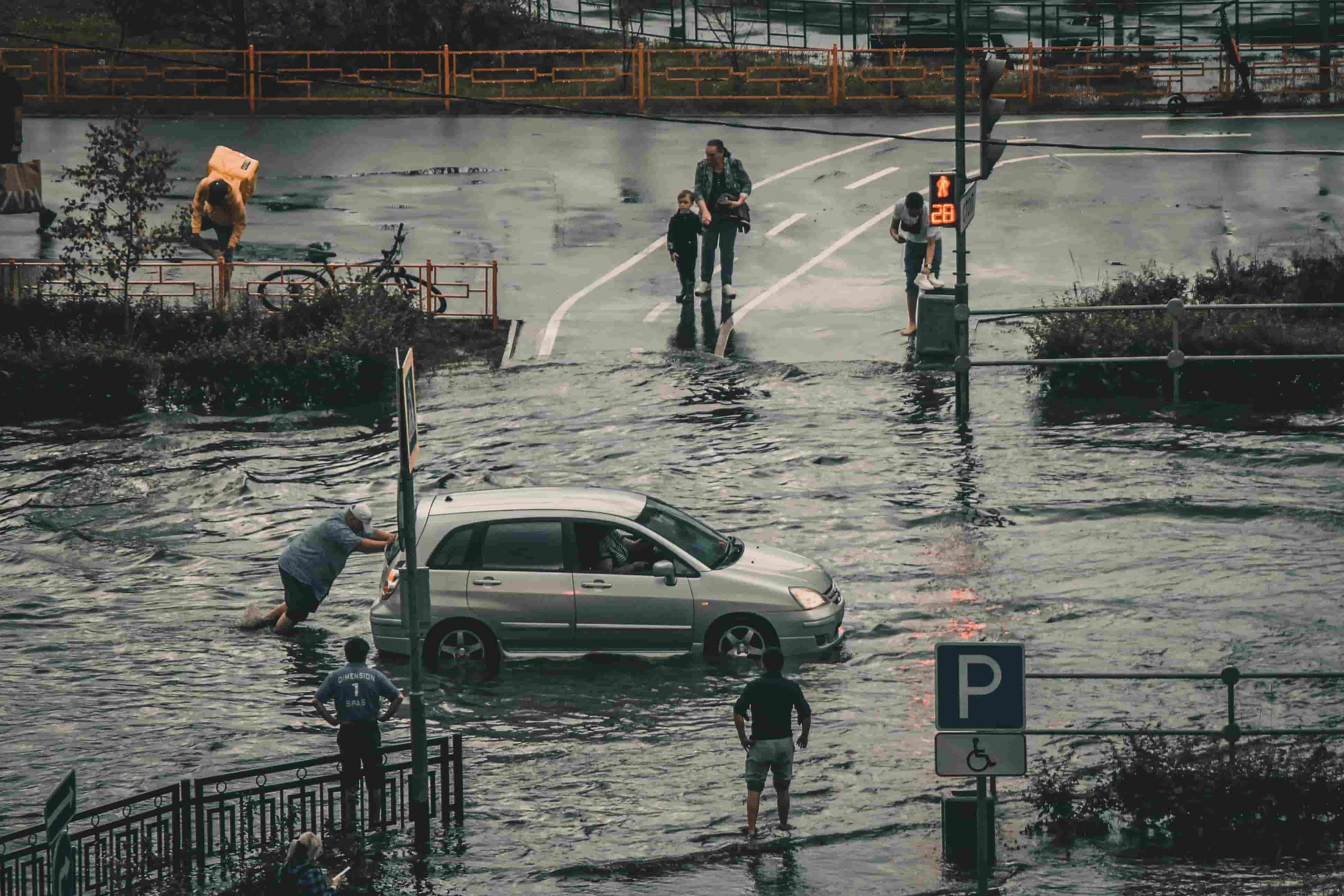 Man pushing a stalled silver hatchback through a flooded street as pedestrians wade across the intersection